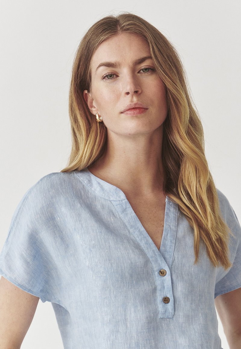 Camicia di lino azzurro chiaro con scollatura a V, maniche corte e due bottoni di legno sul collo. Tessuto morbido e vestibilità rilassata.