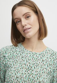 Bloes met bloemenpatroon en een lichte achtergrond met groene en teal bladeren, ronde halslijn en zachte stof met een subtiele textuur.