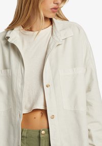 Light beige button-up shirt with two front pockets, made of a textured fabric. Worn over a cream crop top, paired with olive green pants.