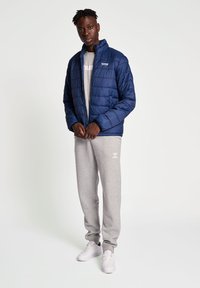 Marinblå vadderad jacka med ståkrage, matchad med gråa sweatpants och vita sneakers. Har en logotyp på vänster bröst.