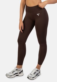 Brązowe legginsy z wysokim stanem i białym logo. Noszone z białymi i czarnymi butami sportowymi, prezentując dopasowany krój.