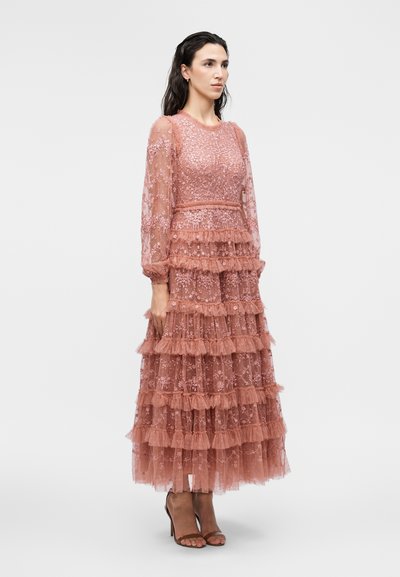 Γυναίκα που φοράει ένα μακρύ, ροζ-σκουριά δαντελένιο φόρεμα με στρώσεις, με κεντήματα λουλουδιών και διαφανή μακριά μανίκια, συνδυασμένο με καφέ πέδιλα με ψηλό τακούνι.