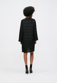 Mulher com cabelo afro, vestindo um vestido preto de renda com bainha em formato de onda até ao joelho e saltos altos pretos com tiras no tornozelo, de pé, virada de costas.