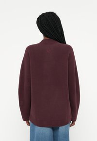 Burgundijska oversized pulover z ribanim robom, detajlom na hrbtni strani ovratnika in diskretno rdečim logotipom. Mehak materiale, dolgi rokavi in sproščen kroj.