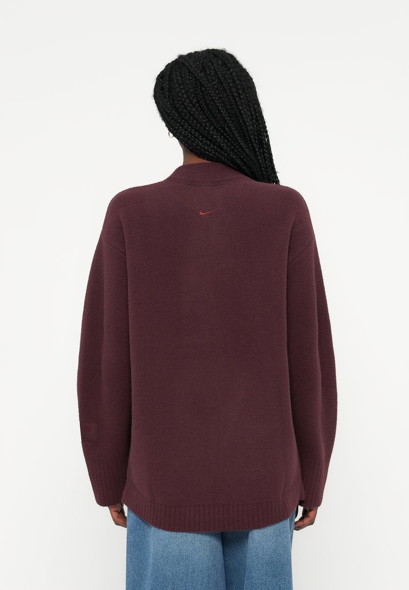 Bordeaux oversized tröja med ribbad fåll, detalj vid bakre halsringning och en subtil röd logotyp. Mjuk textur, långa ärmar och avslappnad passform.