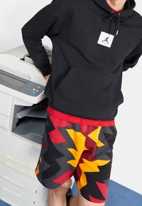 Persona che indossa una felpa nera con cappuccio e logo bianco e pantaloncini colorati a motivo a zigzag, in piedi accanto a una fotocopiatrice su un pavimento blu.