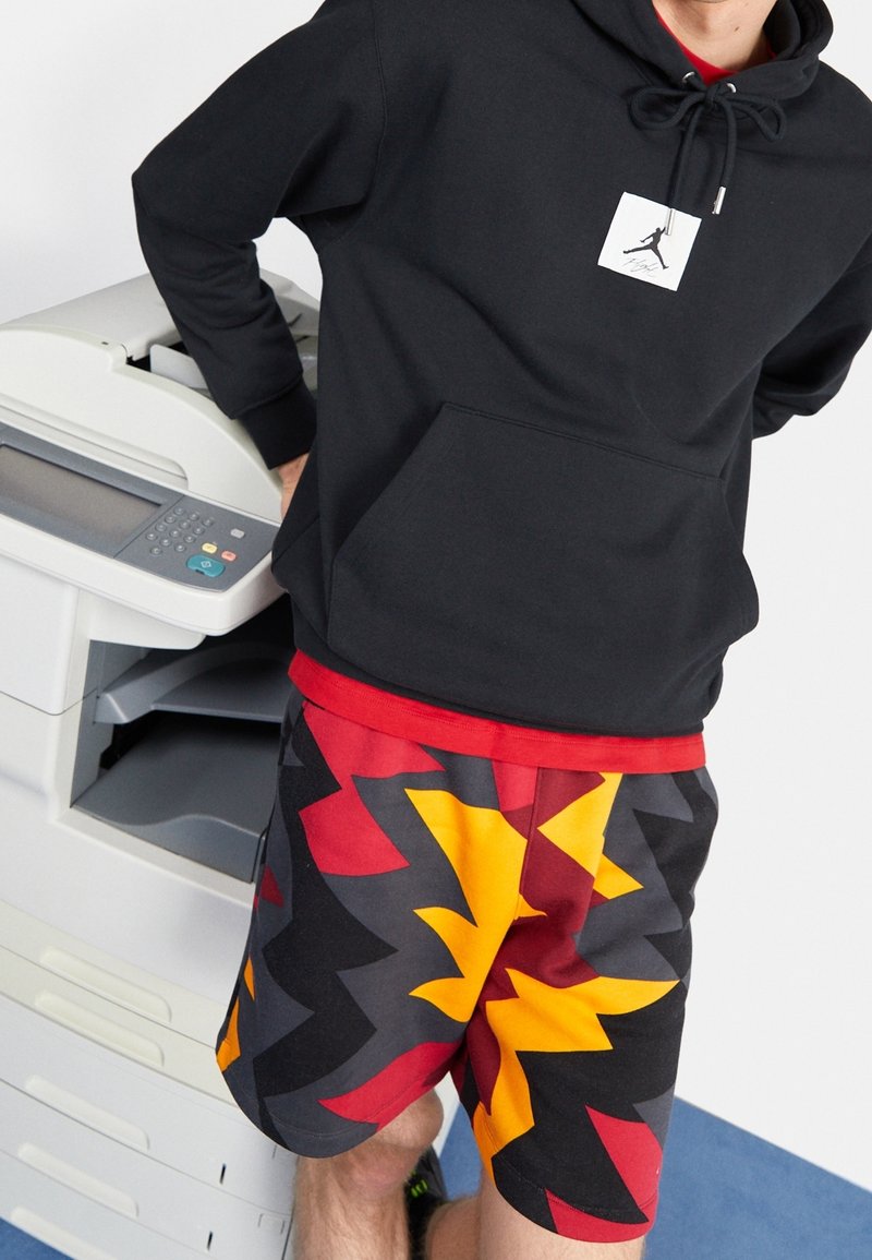 Persona che indossa una felpa nera con cappuccio e logo bianco e pantaloncini colorati a motivo a zigzag, in piedi accanto a una fotocopiatrice su un pavimento blu.