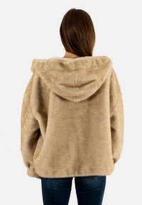 Giacca con cappuccio in pelliccia sintetica beige, con silhouette oversize, texture morbida e orlo arrotondato. La vista posteriore mostra una vestibilità rilassata e un design casual.