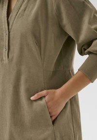 Khaki corduroy tunic with a V-neck, two side pockets, and button detailing. Features long, ribbed sleeves and a relaxed fit.