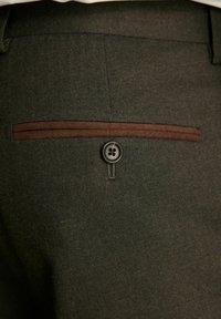 Los pantalones de lana verde oscuro presentan un patrón de espiga, costuras de acento en marrón y un cierre decorativo con botón en el bolsillo trasero.