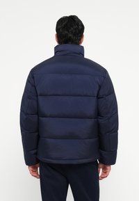 Marineblaue Steppjacke mit hohem Kragen, gepolsterten Abschnitten und glatter, matter Oberfläche. Die Rückansicht zeigt eine lockere Passform und einen geraden Saum.