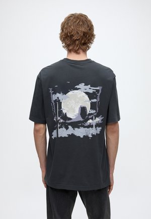 Person mit lockigem Haar, die ein schwarzes T-Shirt mit einem großen weißen Mond und einer Wüstenlandschaft mit Kakteen auf dem Rücken trägt.