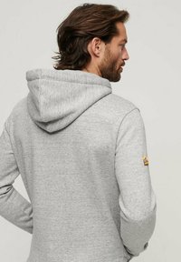 Felpa grigia realizzata in materiale morbido, con un grande cappuccio, maniche lunghe e un piccolo logo accentato sulla manica destra.
