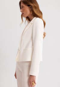 Biała marynarka o dopasowanym kroju, z szpiczastymi klapami i dwoma przednimi kieszeniami. Gładki materiał, minimalistyczny design, w komplecie z pasującymi spodniami.