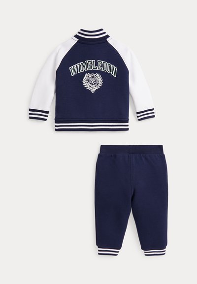 Chándal azul marino con mangas blancas. La chaqueta presenta el logo "WIMBLEDON" y puños a rayas. Los pantalones son de un azul marino sólido con tobillos a rayas.