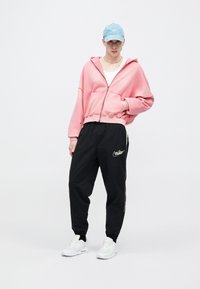 Ροζ φούτερ με φερμουάρ και χαλαρή εφαρμογή, μαύρο παντελόνι τζόγκερ με λευκό λογότυπο Nike και λευκά αθλητικά παπούτσια. Ανοιχτό μπλε καπέλο.