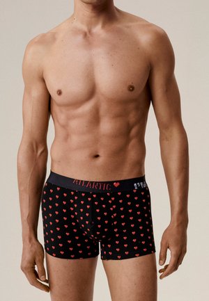 Szczupły męski tors ubrany w czarne bokserki z czerwonym wzorem serca i paskiem oznaczonym napisem "ATLANTIC" na neutralnym tle.