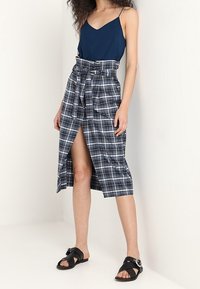 Laivastonsininen toppi yhdistettynä korkeavyötäröiseen ruudulliseen hameeseen, jossa on solmittava vyö ja halkio edessä, sekä mustat tasapohjaiset sandaalit koristeellisilla yksityiskohdilla.