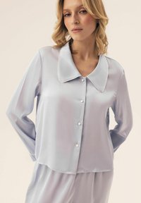 Jasnofioletowa satynowa bluzka piżamy z długimi rękawami, dużym kołnierzem Peter Pan i białymi guzikami. Gładka tekstura i luźny krój.