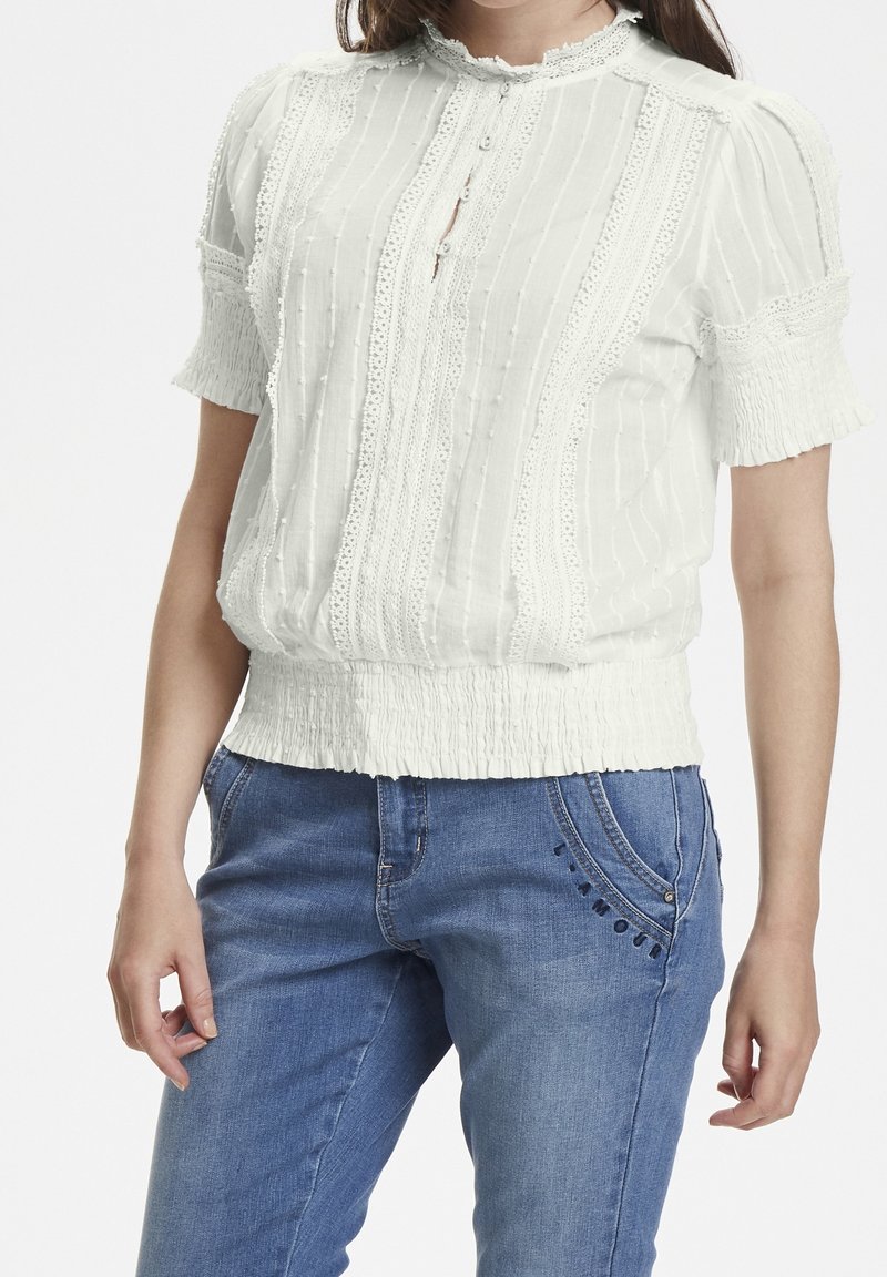 Blusa bianca tessuta con decorazioni in pizzo, maniche corte e orlo arricciato. Abbinata a jeans in denim blu con cuciture decorative.