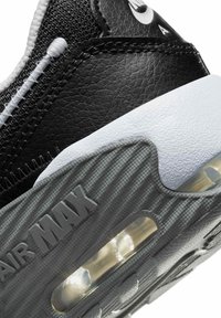 Zapatilla sintética negra con acentos blancos, que presenta una superficie texturizada, una cámara de aire visible y un logo "AIR MAX" en relieve en el lateral.