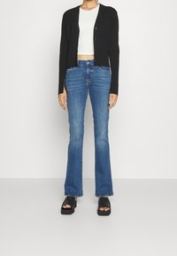 Person iført en sort knaplukket cardigan, hvid crop top, blå vide jeans og sorte platformsandaler, der står foran en ensfarvet baggrund.