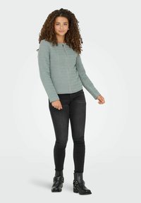 Ung kvinna med lockigt hår som bär en ljusgrå zip-up jacka, svarta tajta jeans och svarta ankelstövlar, stående mot en enkel bakgrund.