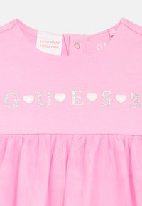 Ροζ φόρεμα με φούστα από πιεσμένο τούλι, με τη λέξη "GUESS" να σχηματίζεται από λαμπερές διακοσμήσεις σε σχήμα καρδιάς στο στήθος.