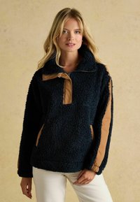 Granatowy polarowy pulower z półzamkiem, beżowymi wstawkami na kołnierzu i rękawach, wykonany z teksturowanego, miękkiego materiału. Z bocznymi kieszeniami.