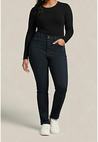Femme portant un haut noir à manches longues, un jean slim foncé et des baskets blanches, debout avec une main dans la poche sur un fond clair.