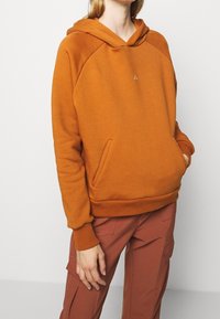 Sudadera naranja con un bolsillo canguro, puños y dobladillo acanalados. Presenta un pequeño logo en el pecho. La tela tiene una textura suave.