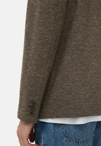 Brun struktureret blazer med detalje i form af tre knapper på manchetterne. Båret over en hvid skjorte og parret med blå denim jeans.