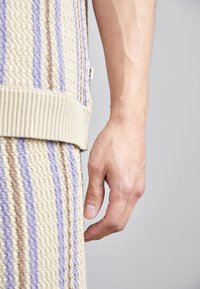 Gebreide trui set met een gestructureerd patroon in beige, paarse en bruine strepen; geribbelde boorden; zichtbare wasinstructies aan de zijkant. Hand rust op de stof.