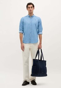 Lichtblauw button-up shirt, crèmekleurige broek en zwarte schoenen. Het model houdt een marineblauwe gestructureerde tote bag vast met meerdere zakken.