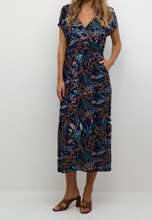Femme portant une robe portefeuille bleu marine à motif floral avec des manches courtes et des sandales marron à talons, debout avec une main dans la poche sur un fond uni.