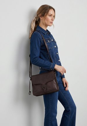 Frau in blauer Jeansjacke und Hose mit hellbraunem Schal im Haar, trägt eine dunkelbraune Lederschultertasche und steht an einer grauen Wand.