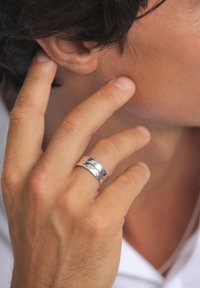 Anello d'argento indossato su una mano che tocca il volto, con una superficie lucida e due fasce distinte, una delle quali incisa con un motivo.