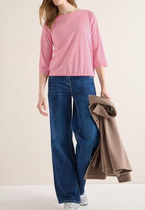 Femme portant un haut rayé rose et blanc, un jean bleu à jambes larges, et des baskets argentées, tenant une veste beige pliée dans sa main droite.