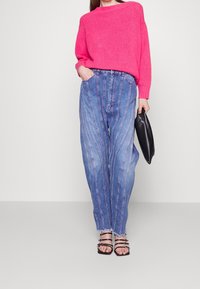 Helles pinkes Strickoberteil, hochtaillierte blaue Jeans mit vertikalen Streifen, fransigem Saum und schwarzen Riemchensandalen, die eine schwarze Clutch halten.