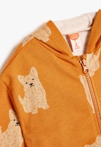 Orange zip-up hoodie til spædbørn med et tekstureret hundemønster i cremefarve. Ribbede kraver og lynlås i gulltone. Blød stofstruktur.