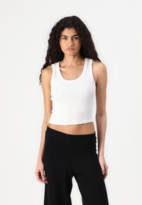 White ribbed tank top with a round neckline and wide straps, cropped length, paired with black loose-fit pants. Clean, minimalist design.