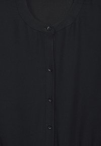 Chemisier noir à boutonner avec un col rond, comportant six boutons noirs sur le devant et une texture de tissu douce et lisse.