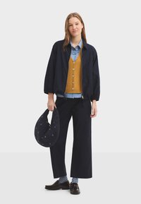 Blouson aviateur marine, gilet en maille moutarde avec boutons, chemise à col bleu clair, pantalon à jambes larges, sac bleu marine à motif, chaussures marron.