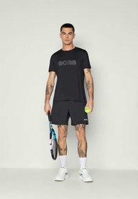 Crna sportska majica s "BORG" logotipom, uparen s crnim kratkim hlačama. Osoba drži tenisku rekete i lopticu, noseći bijele tenisice.