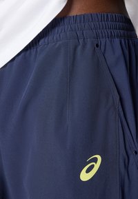 Ναυτικό μπλε αθλητικά σορτς με ελαστική μέση και κίτρινο λογότυπο Asics κοντά στην δεξιά τσέπη, φορεμένα με λευκό μπλουζάκι.