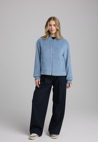 Lichtblauwe fleece bomberjack met zijzakken en geribde manchetten, gecombineerd met zwarte wijde broeken en beige sneakers. Soepele textuur.