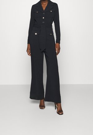 Black jumpsuit with long sleeves, buttoned front, chest pockets, and waist tie. Wide legs and smooth fabric with a soft texture.