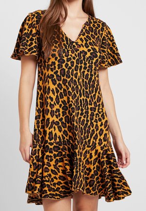 Femme portant une robe à manches courtes et col en V, avec un imprimé léopard orange et noir et un ourlet volanté, se tenant devant un arrière-plan clair uni.