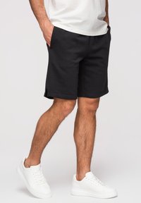 Homme portant un short noir jusqu'aux genoux, des baskets blanches décontractées et un t-shirt blanc, debout les mains dans les poches devant un fond uni.
