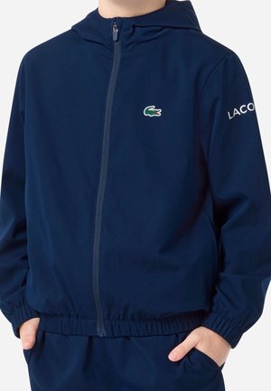 Chłopiec ubrany w granatową kurtkę na zamek z kapturem, z elastycznymi mankietami i pasem, z logo Lacoste na piersi i rękawie, z rękami w kieszeniach.